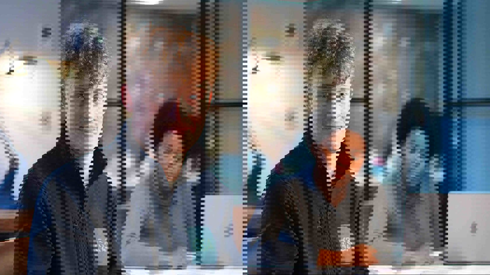 A young man with curly red hair and a navy jacket featuring the "GSA" logo, seated in an office, smiling at the camera. Another person with dark hair is visible at a table in the background.