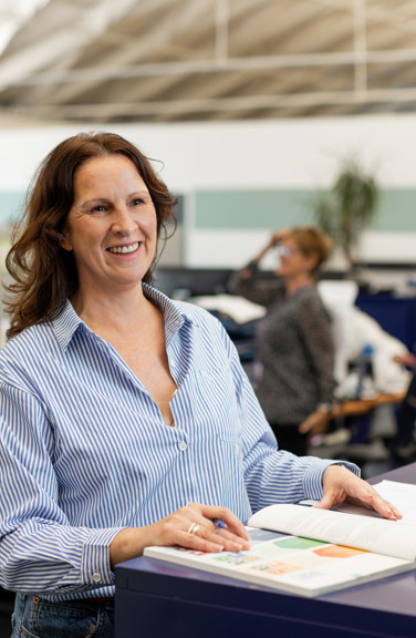 A person smiling and wearing a blue blouse stands at a table looking at finance reports