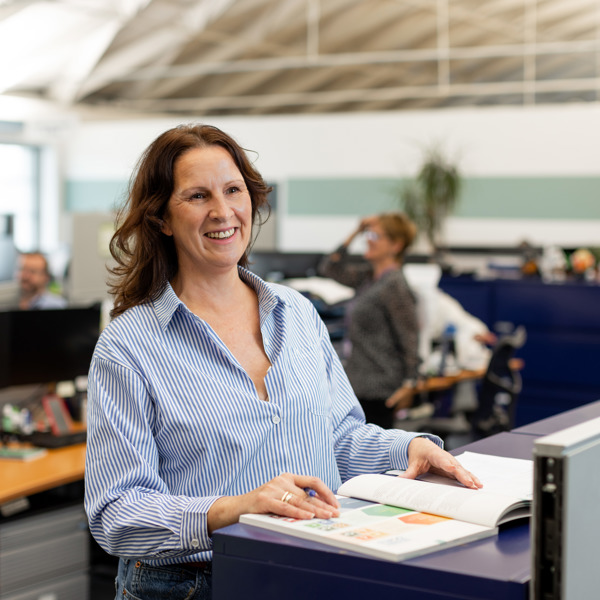 A person smiling and wearing a blue blouse stands at a table looking at finance reports