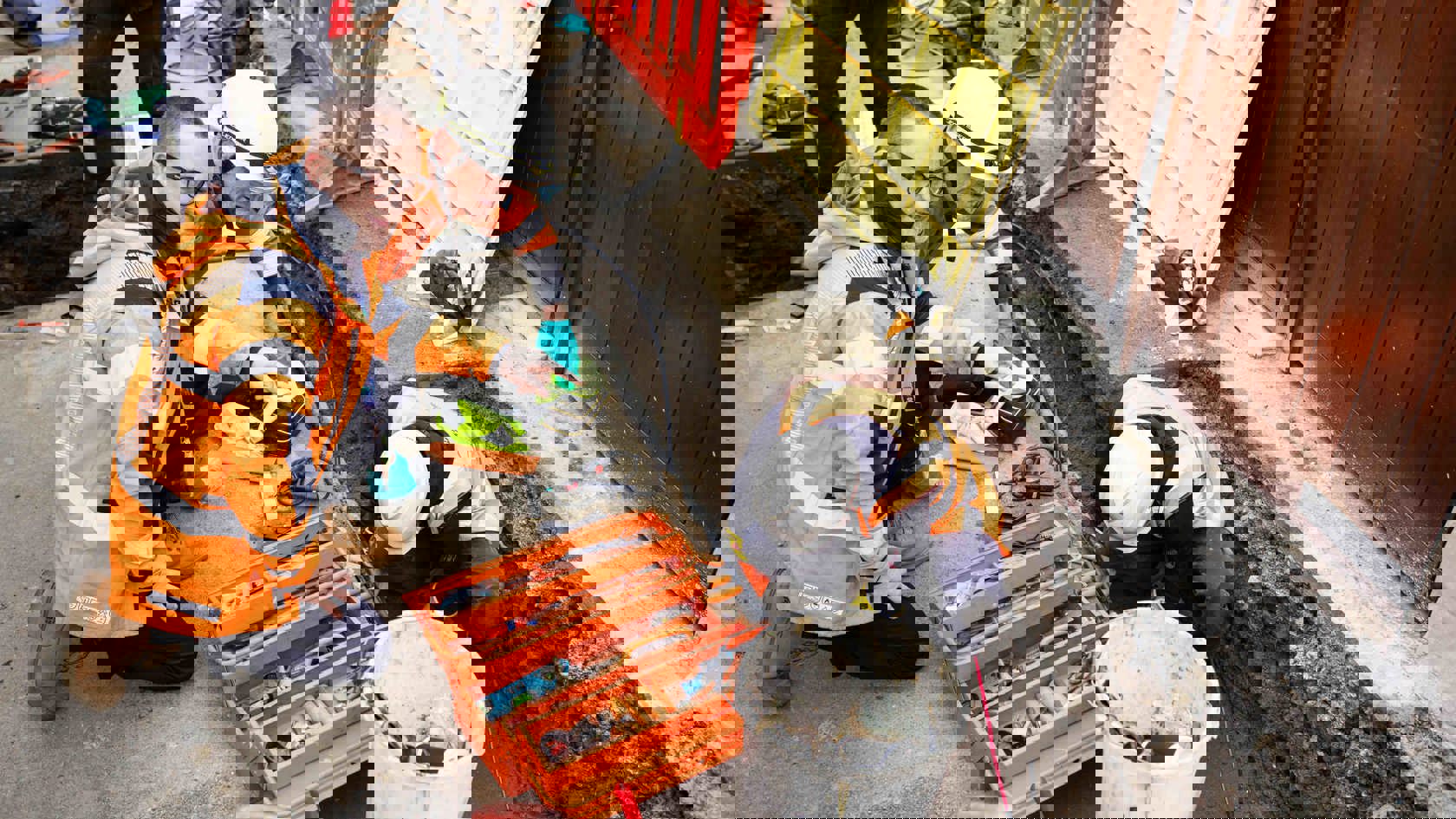 Two people wearing high vis jackets crouched down, looking into an open roadworks pit. There's a person in the pit wearing high vis and a hard hat