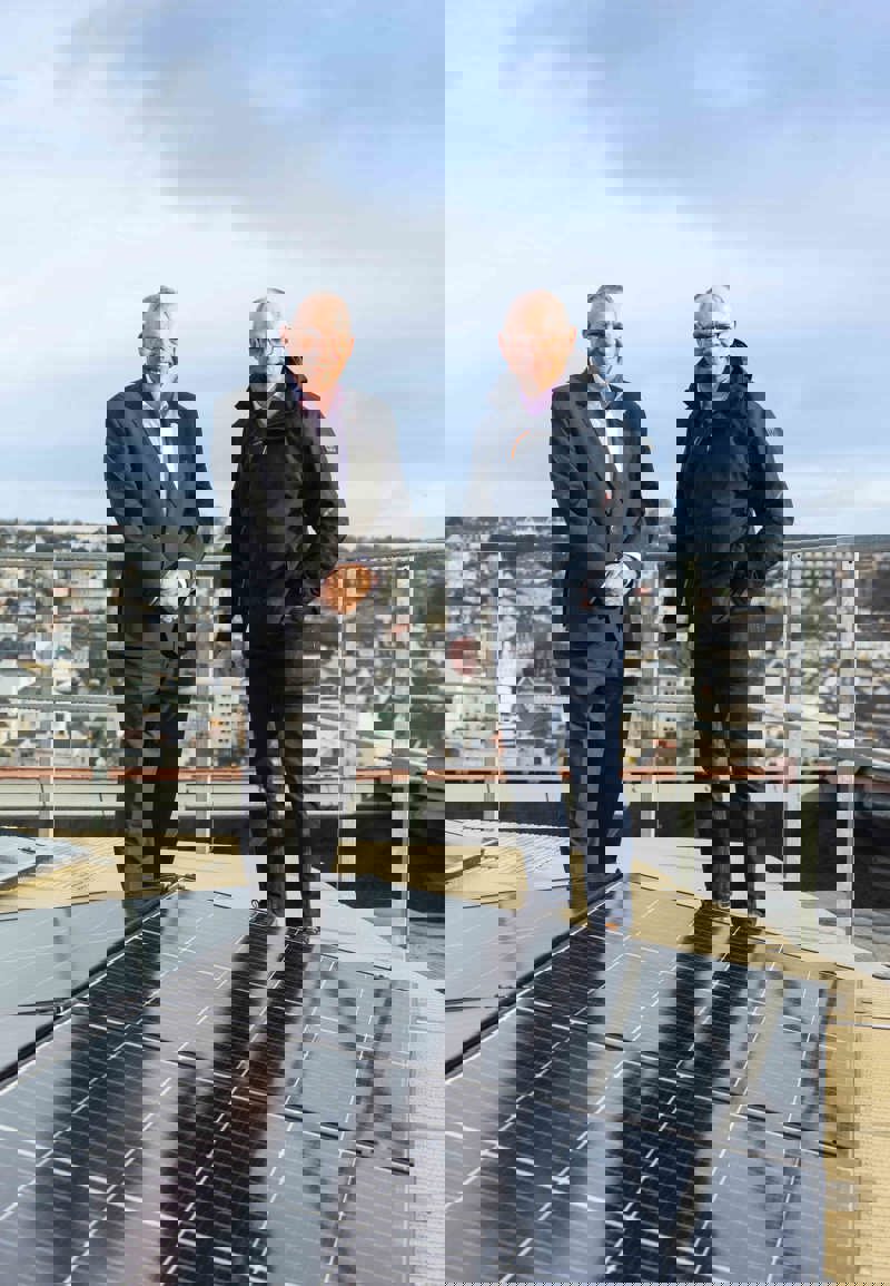 Two people dressed in suits standing on a rooftop next to solar PV panels. There are town buildings in the background.