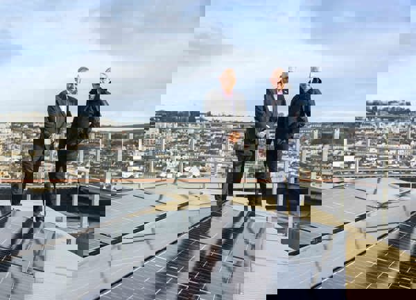 Two people dressed in suits standing on a rooftop next to solar PV panels. There are town buildings in the background.