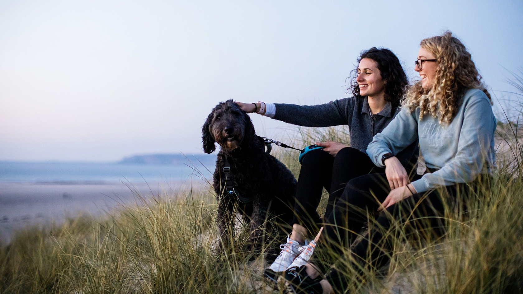 Two female friends sit on the sand dunes at St Ouens, the girl on the left pats a black dog.
