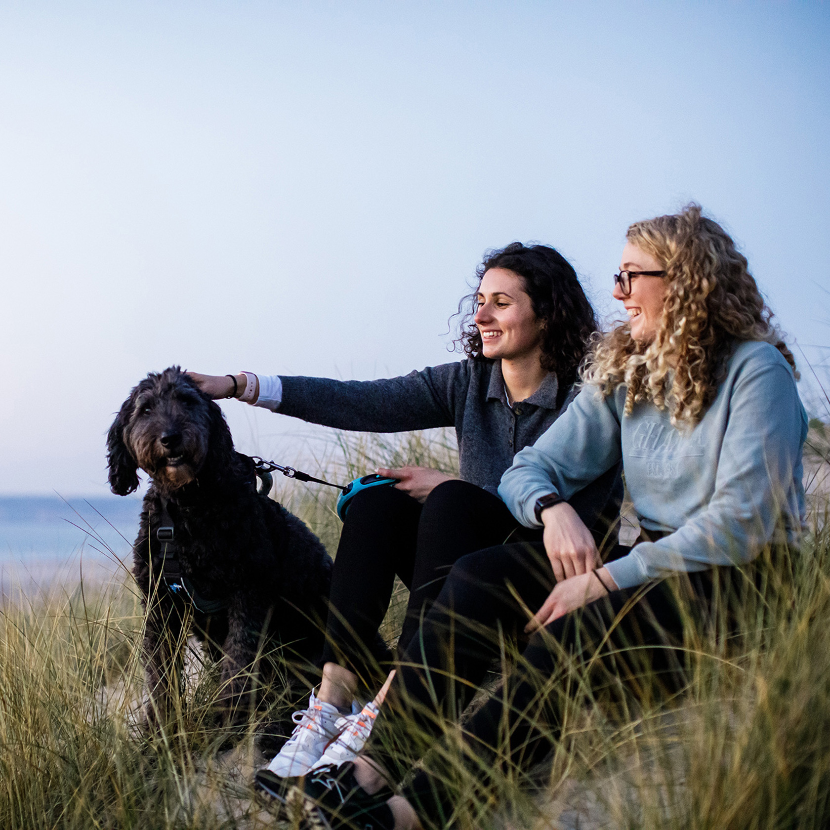 Two female friends sit on the sand dunes at St Ouens, the girl on the left pats a black dog.