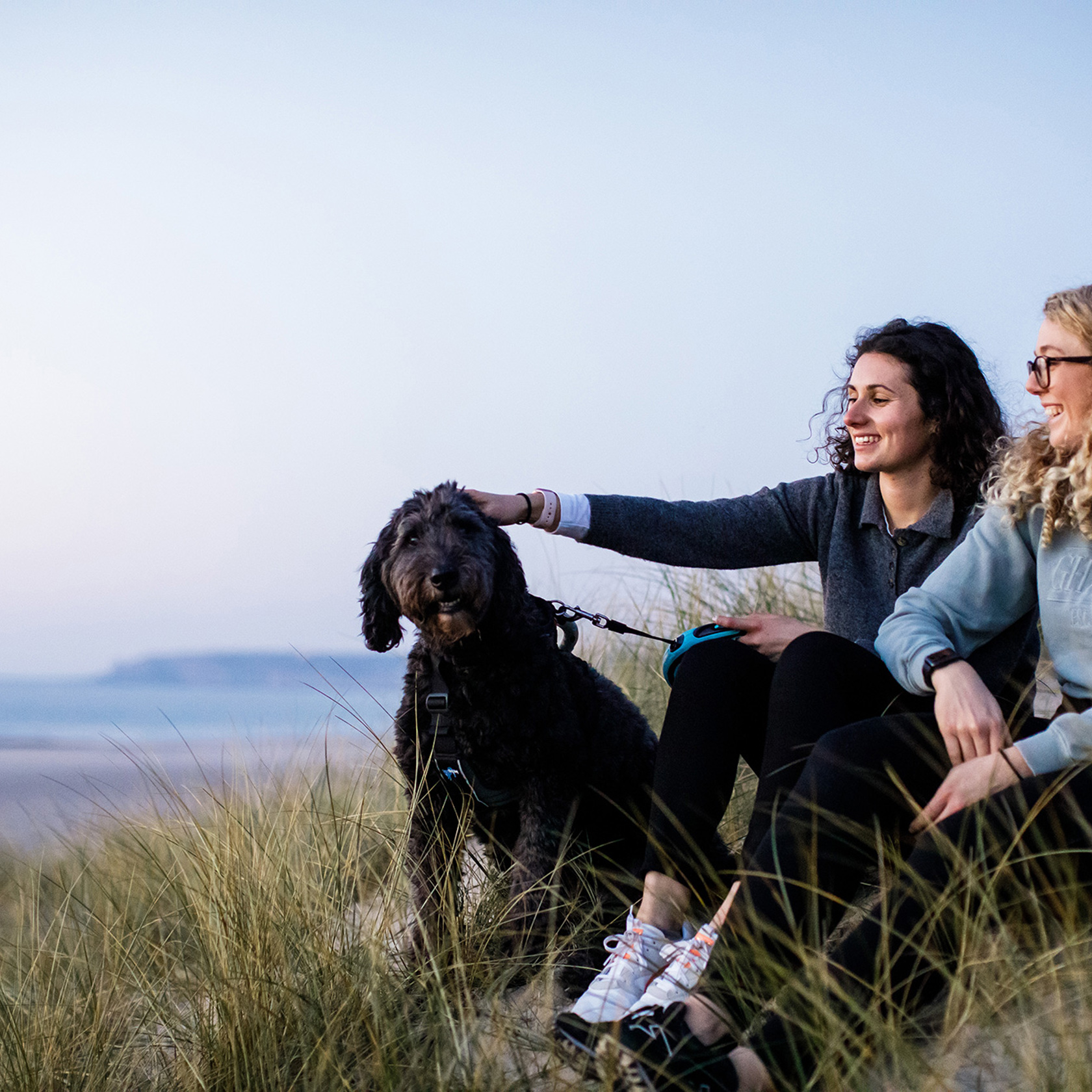 Two female friends sit on the sand dunes at St Ouens, the girl on the left pats a black dog.