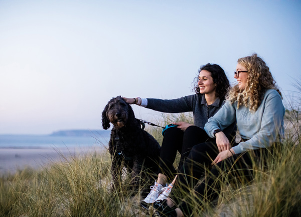 Two female friends sit on the sand dunes at St Ouens, the girl on the left pats a black dog.