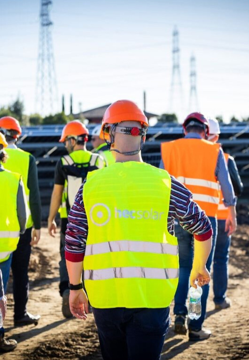 A group of people in high vis jackets walking through a field with solar panels. 