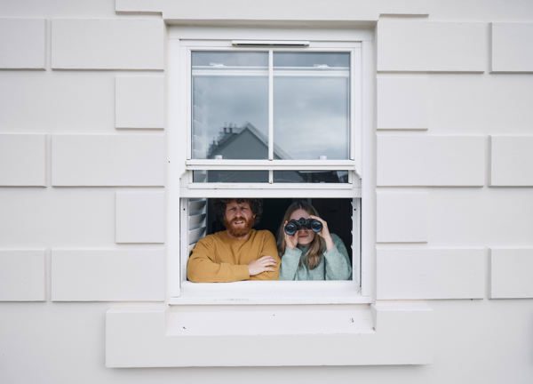 A man and a woman looking out of the upstairs windown of their house, the woman is using binocculars.