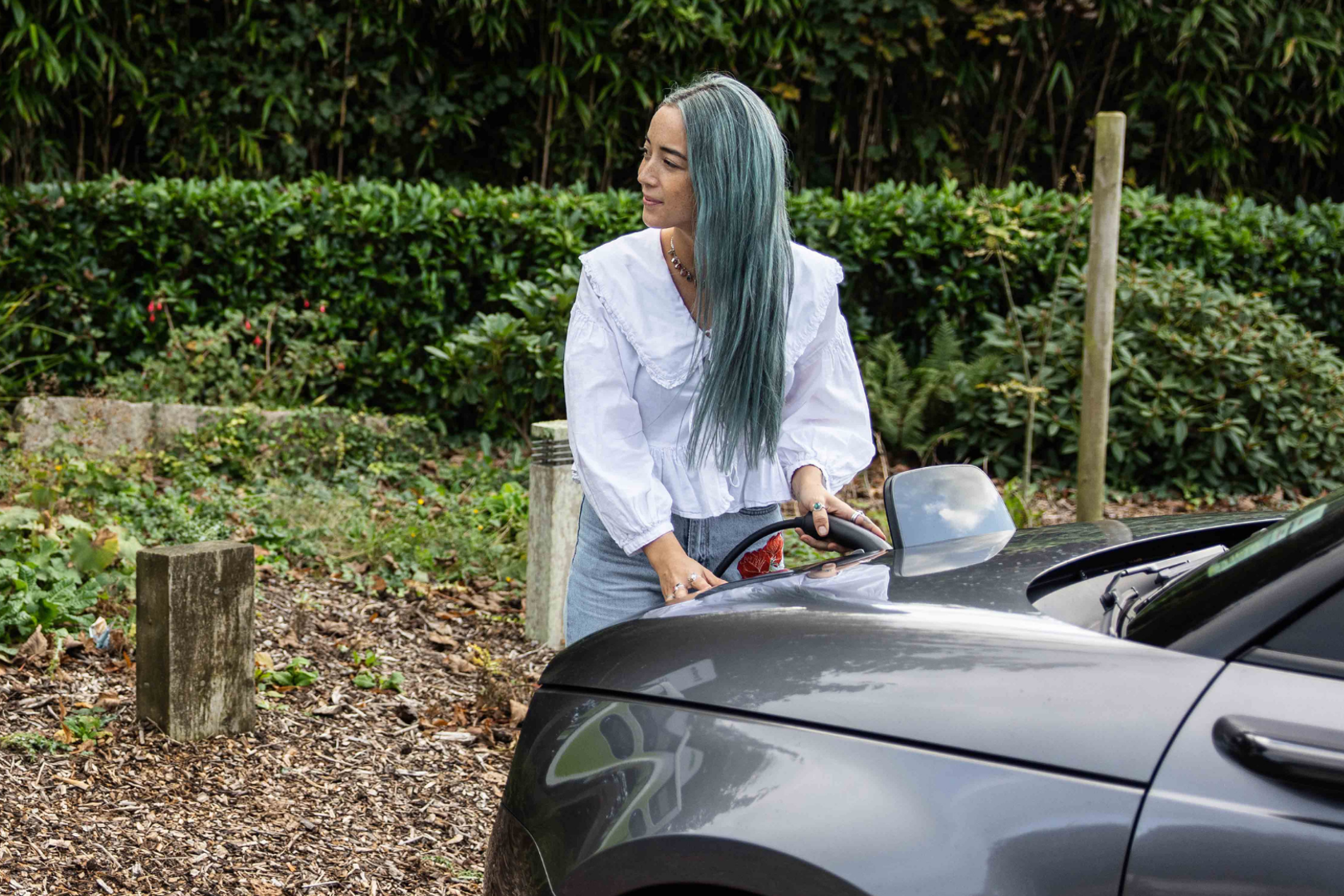Woman with teal hair wearing a white ruffled blouse and denim jeans charges a grey electric car in an outdoor setting with lush greenery.