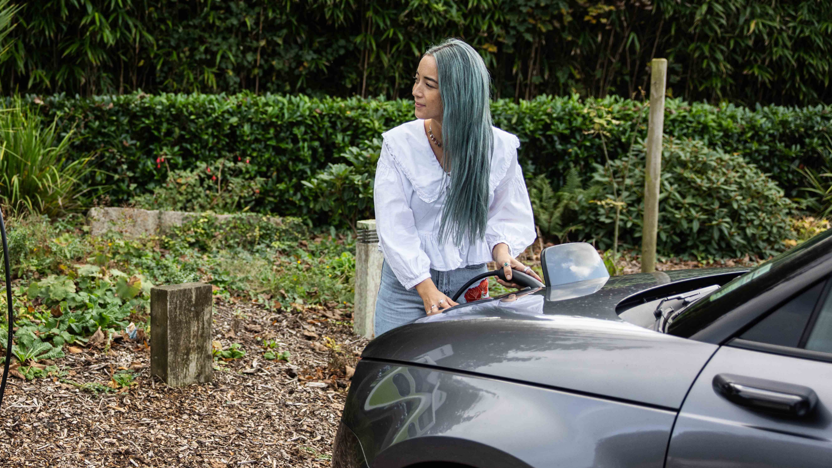 Woman with teal hair wearing a white ruffled blouse and denim jeans charges a grey electric car in an outdoor setting with lush greenery.