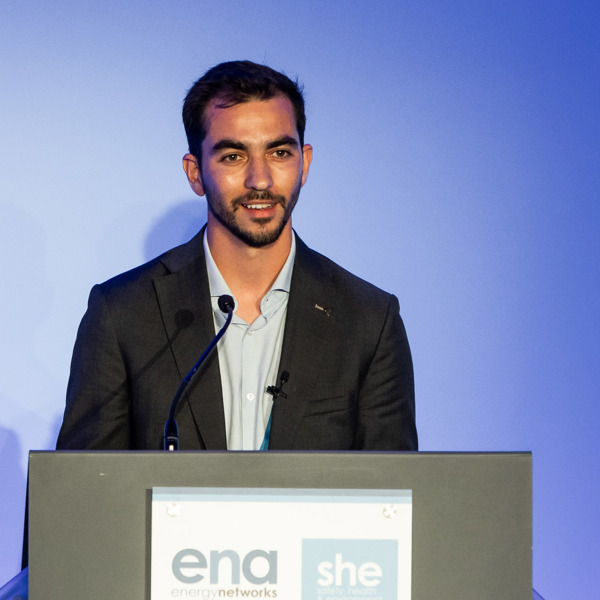 Tito Ornelas wearing a shirt and jacket, standing in front of a blue background and behind a podium giving a talk at a conference