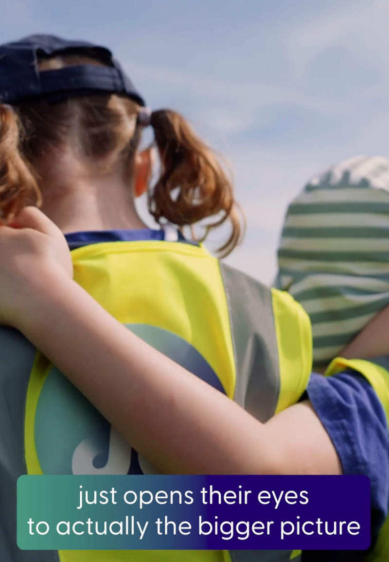 A photo showing the backs of a group of primary school children walking through a solar farm. The children have their arms around each other's shoulders and are wearing yellow high vis vests with the  JE logo on the back