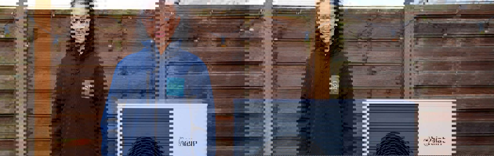 A woman in a blue GSA jacket and black pants stands in a patio next to a Vaillant aroTHERM plus heat pump. She smiles and faces the camera.