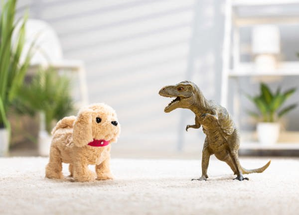 Golden fluffy toy dog wearing a red collar standing next to a plastic toy dinosaur on cream carpet.