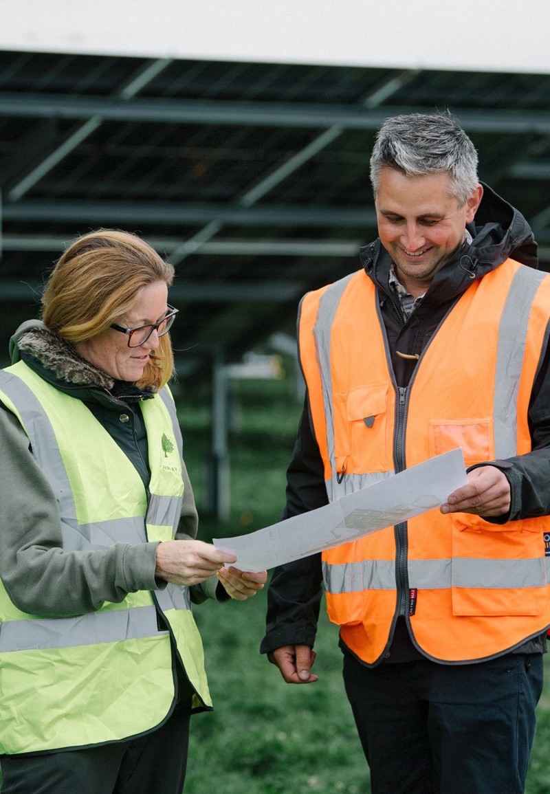 Two people standing in a field with ground mount solar panels behind them, wearing high vis jackets and looking at a piece of paper