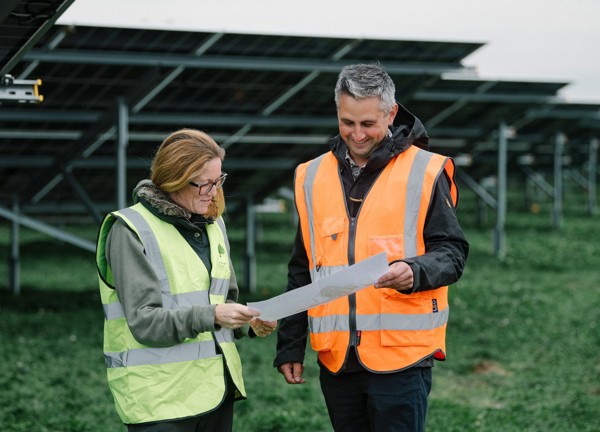 Two people standing in a field with ground mount solar panels behind them, wearing high vis jackets and looking at a piece of paper