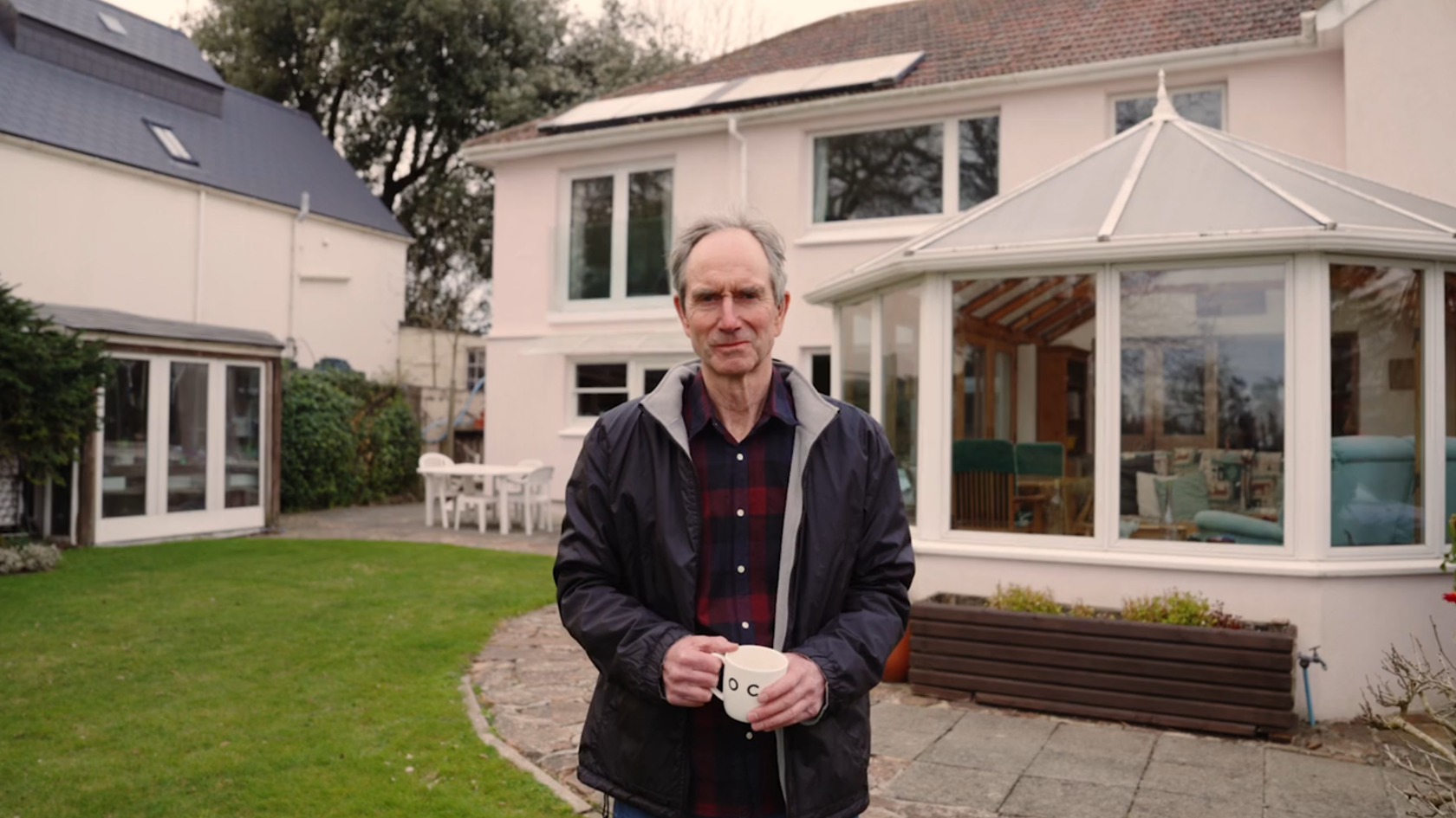 Peter standing in his back garden, wearing glasses, a navy rain jacket and holding a white mug.