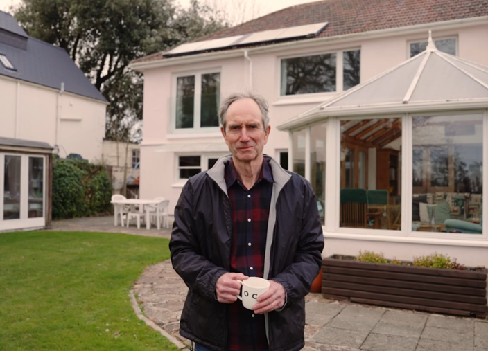 Peter standing in his back garden, wearing glasses, a navy rain jacket and holding a white mug.