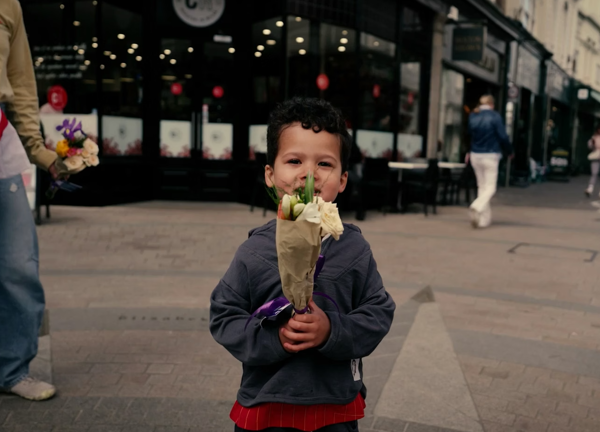A young boy smiling, holings a small bunch of flowers to his face. Shops and passers by are in the background