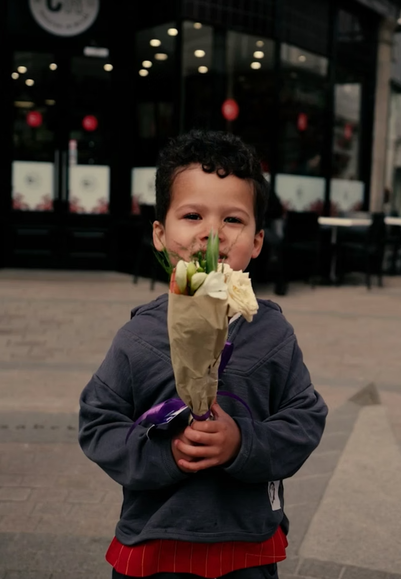 A young boy smiling, holings a small bunch of flowers to his face. Shops and passers by are in the background