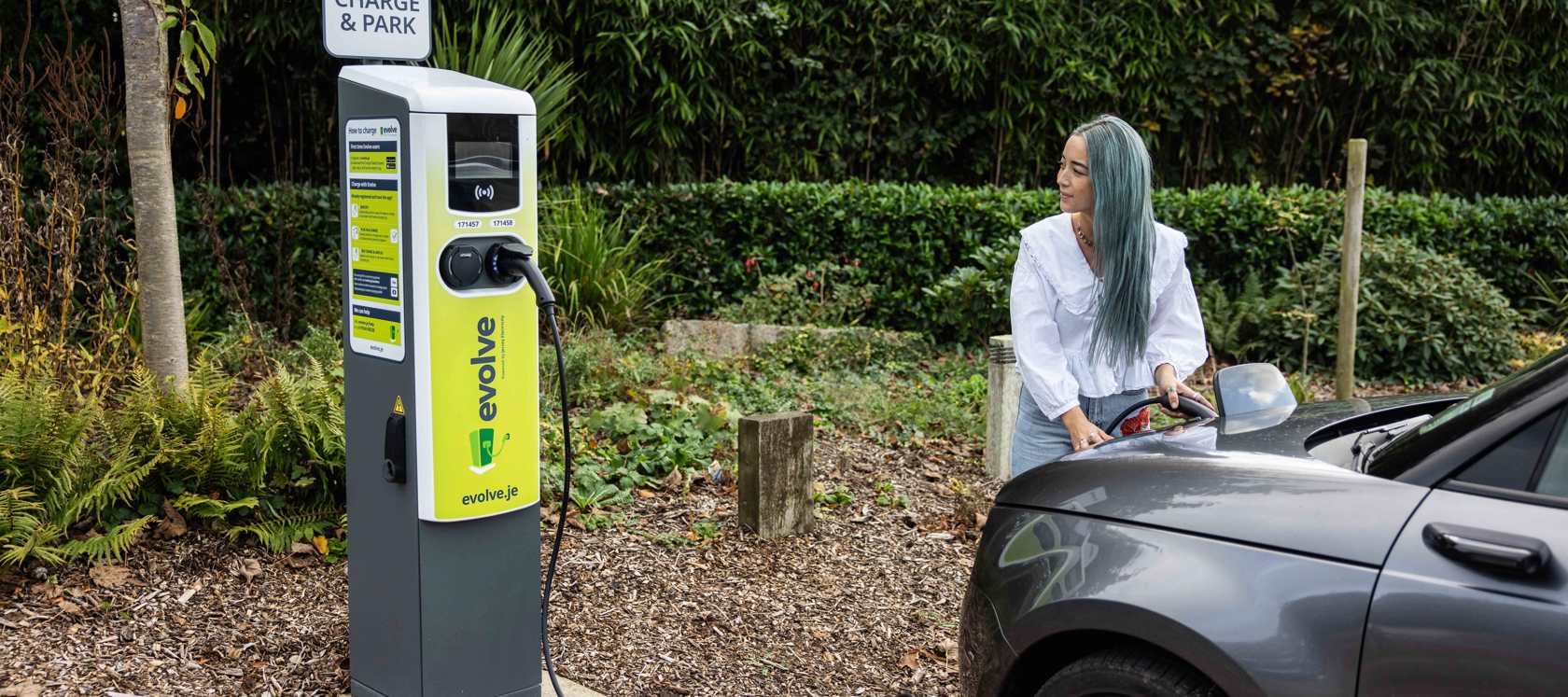 A person with blue-tinted hair connects a charging cable from an EV charger to a dark gray electric car.
