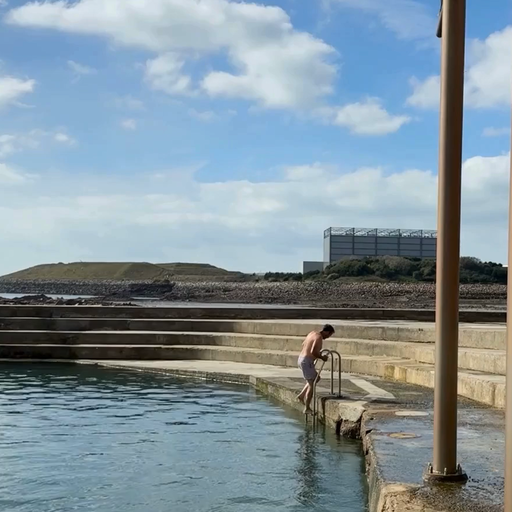 A person in swimming shorts stepping backwards down a pool ladder into the sea. La Collette and the Energy to Waste plant can be seen in the background