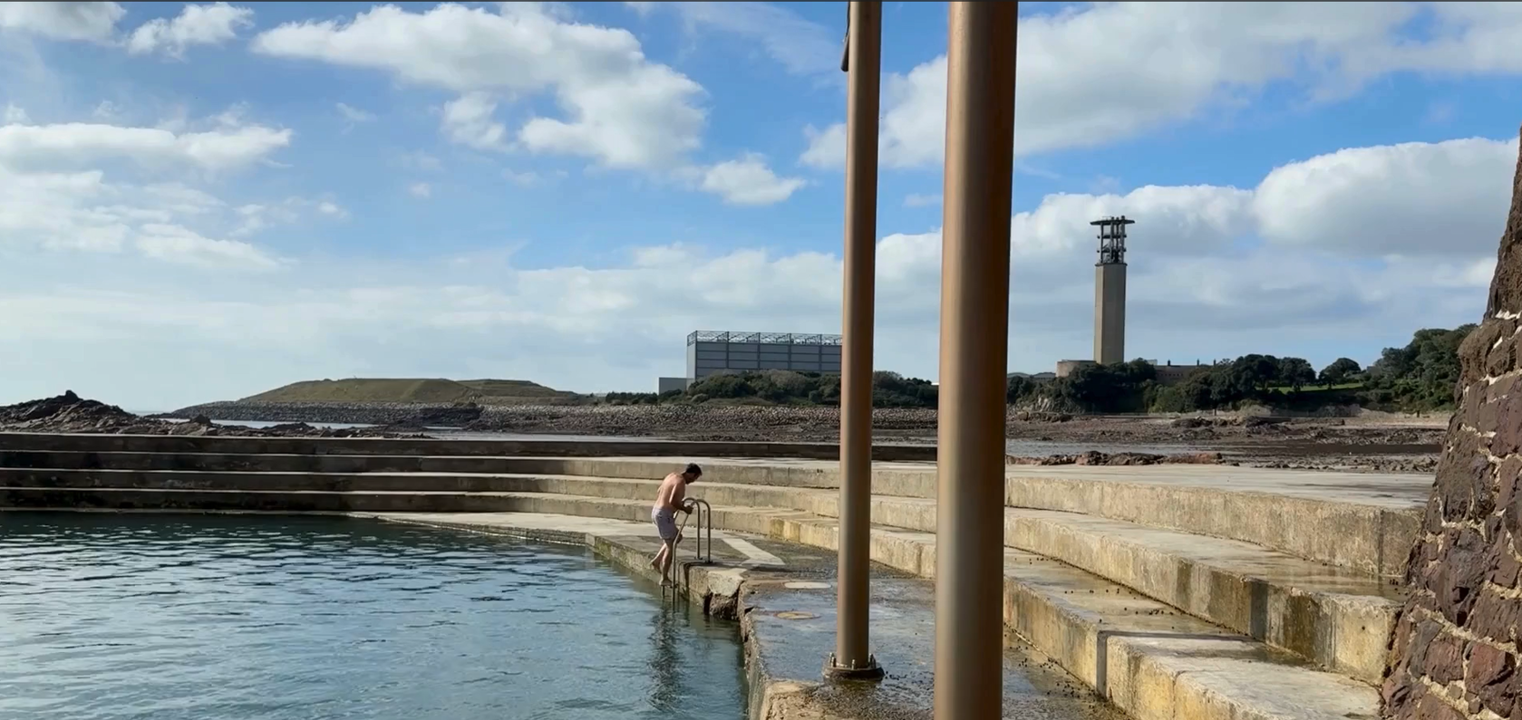 A person in swimming shorts stepping backwards down a pool ladder into the sea. La Collette and the Energy to Waste plant can be seen in the background
