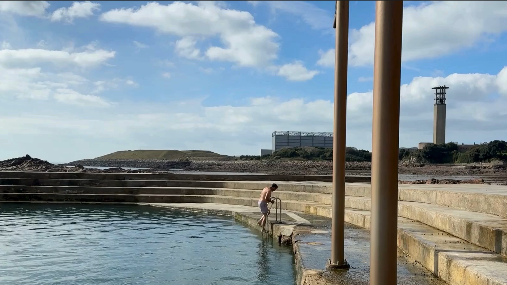 A person in swimming shorts stepping backwards down a pool ladder into the sea. La Collette and the Energy to Waste plant can be seen in the background