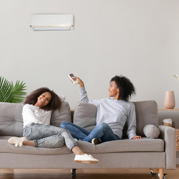 A mother and daughter sit on a couch in a modern home. The mother is operating her air conditioning unit with the remote.