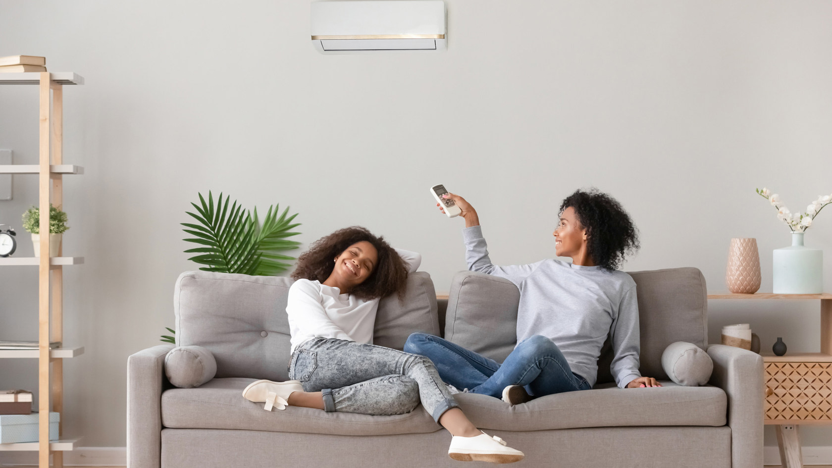 A mother and daughter sit on a couch in a modern home. The mother is operating her air conditioning unit with the remote.