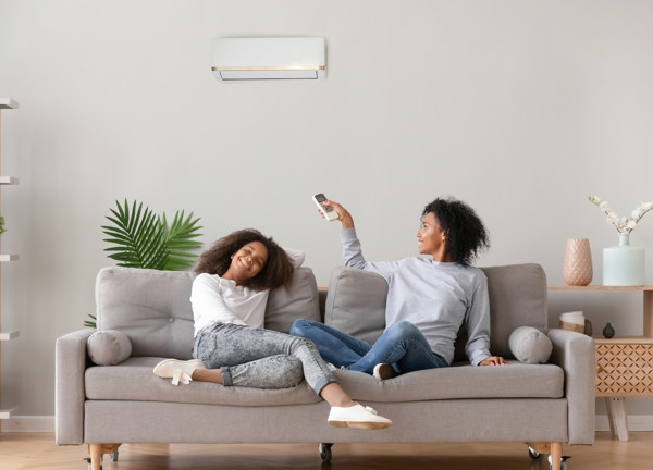 A mother and daughter sit on a couch in a modern home. The mother is operating her air conditioning unit with the remote.