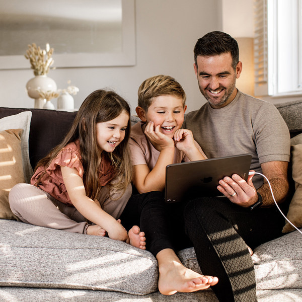 A father and his children enjoy watching a tablet whilst sitting on a comfortable sofa.