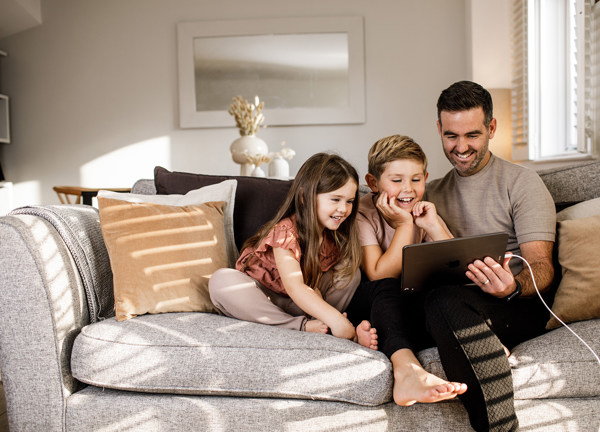 A father and his children enjoy watching a tablet whilst sitting on a comfortable sofa.