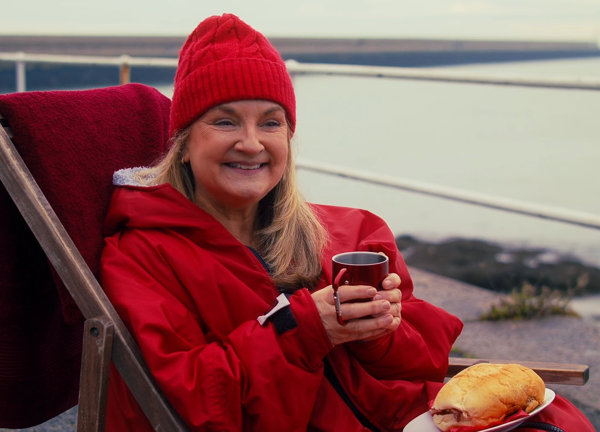 Woman in red beanie and jacket seated in a deckchair, holding a red mug and smiling, with a sandwich on a plate; sea and white railing in the background.