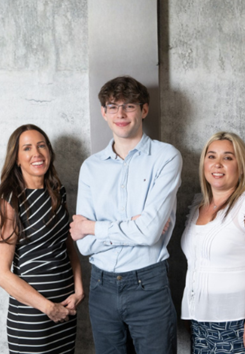 Four people stand together in front of a textured gray wall. A woman in a black and white striped dress stands on the far left, followed by a young man in a light blue button-down shirt, a woman in a white ruffled top, and a man in a tan button-down shirt over a white t-shirt on the far right.