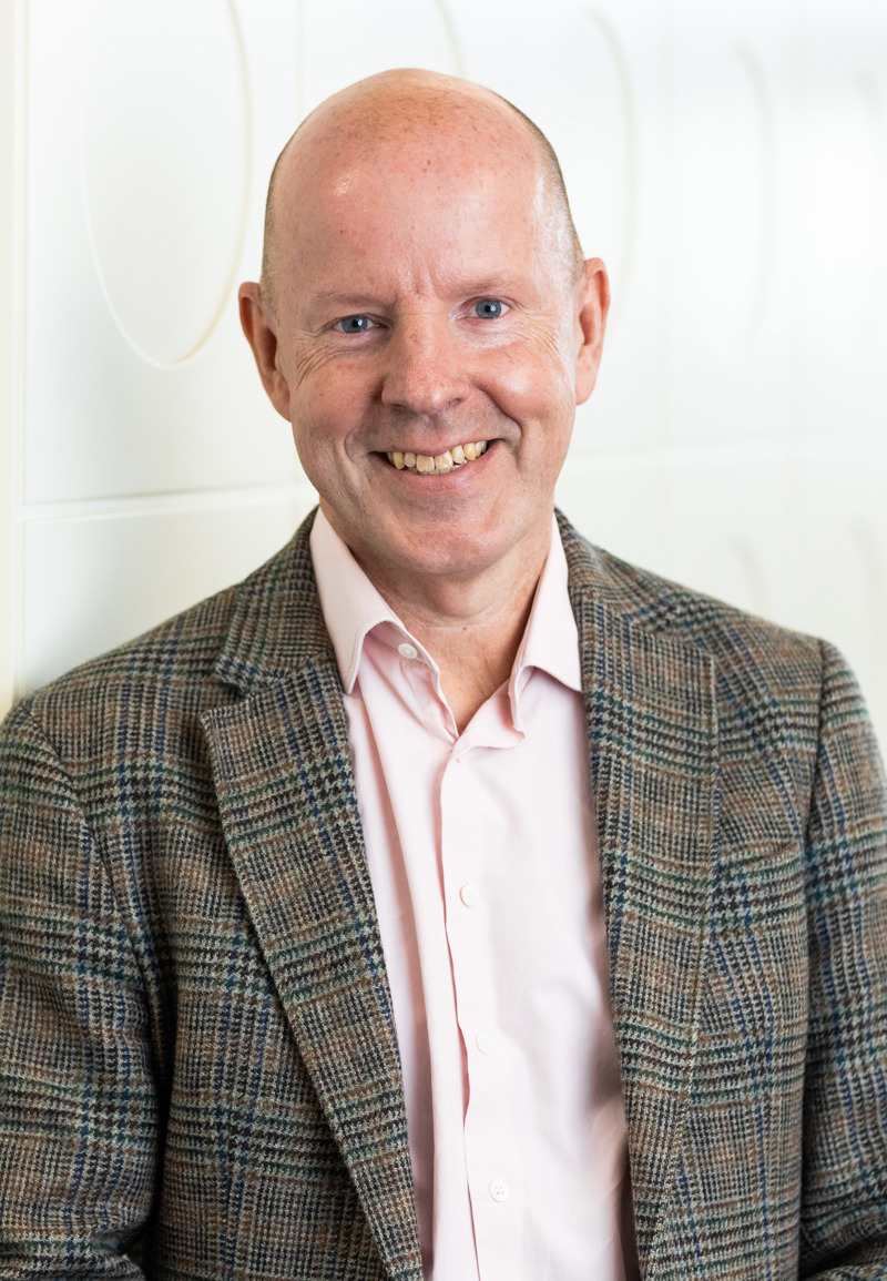 A smiling person standing against a white wall wearing a pink shirt and grey checked blazer.