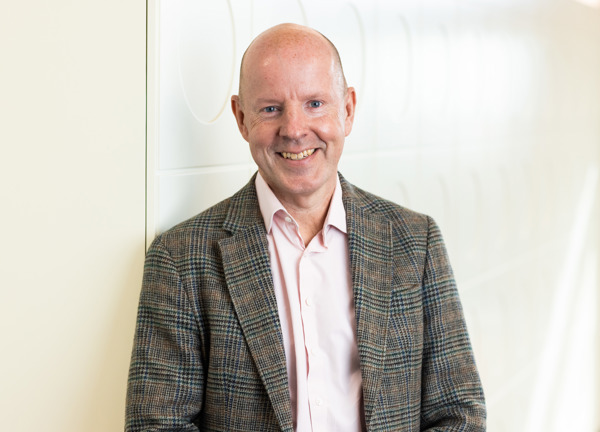 A smiling person standing against a white wall wearing a pink shirt and grey checked blazer.