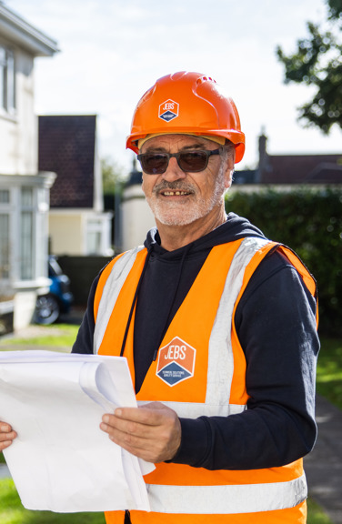 Two construction workers in orange safety vests and helmets inspect the exterior of a house; one holds papers and smiles at the camera, while the other examines a window.