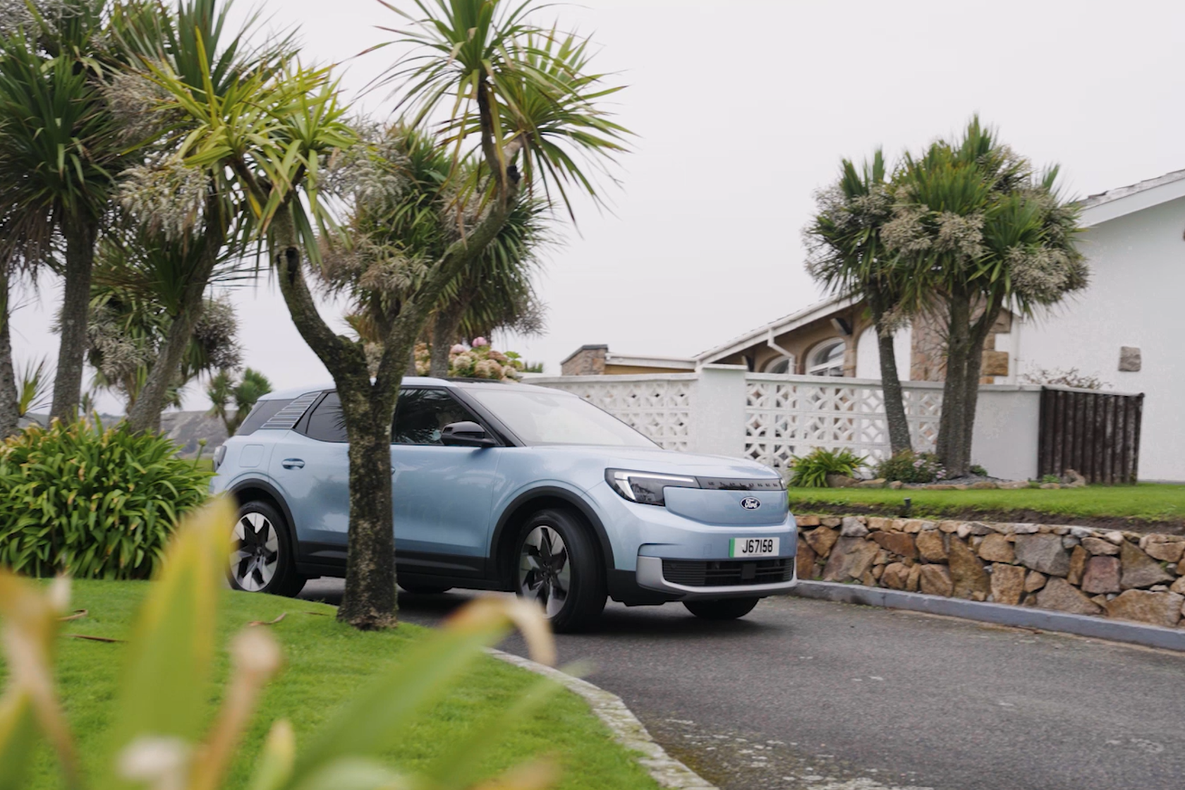 A light blue electice Ford Explorer pulling into a drivewhich has grass and palm trees either side. A house sits in the backgroud