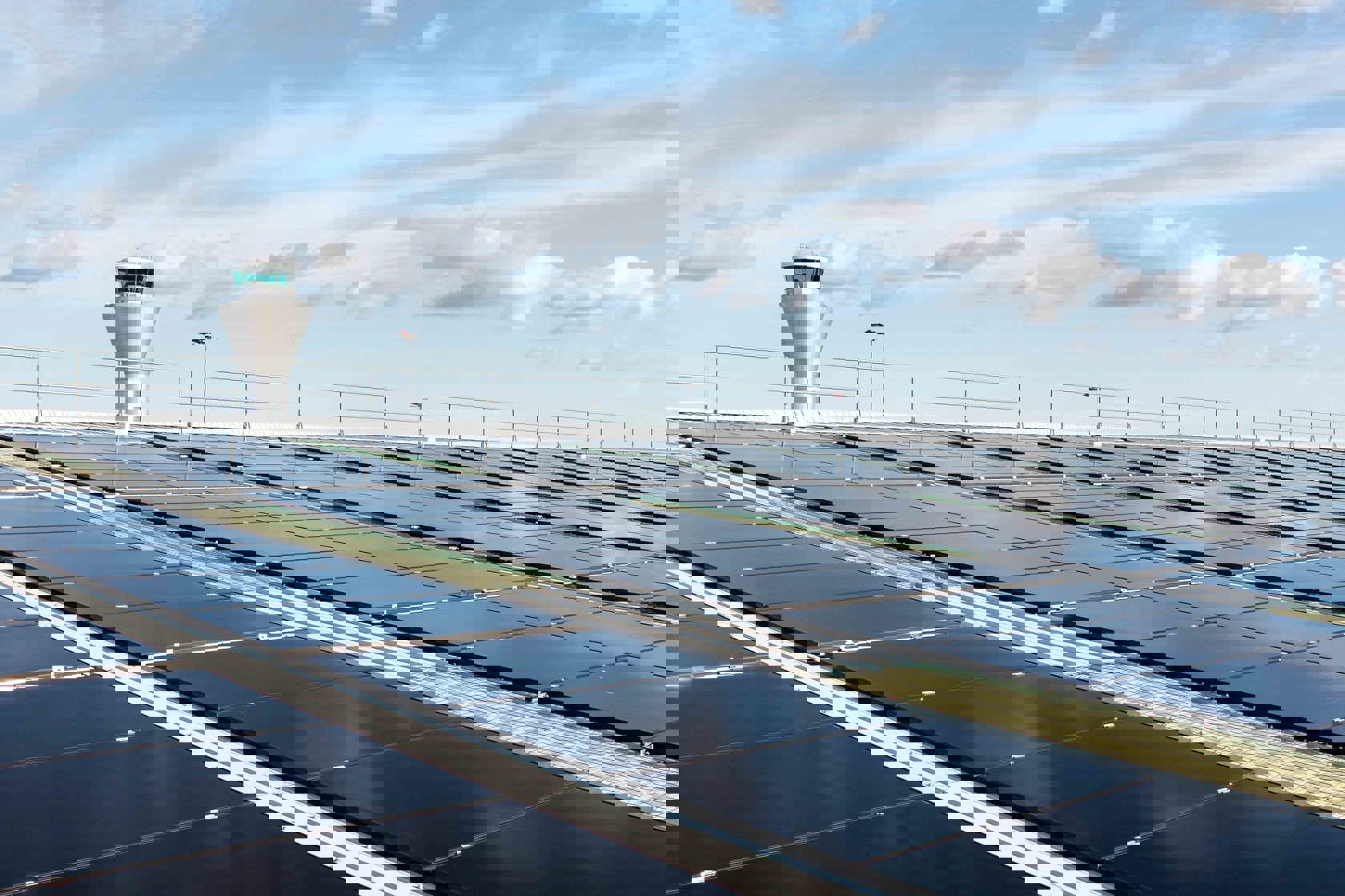 Rows of black solar panels installed on a large rooftop under a bright blue sky with scattered clouds. In the background, an airport control tower is visible, indicating the location near an airport facility.