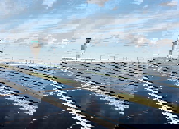 Rows of black solar panels installed on a large rooftop under a bright blue sky with scattered clouds. In the background, an airport control tower is visible, indicating the location near an airport facility.