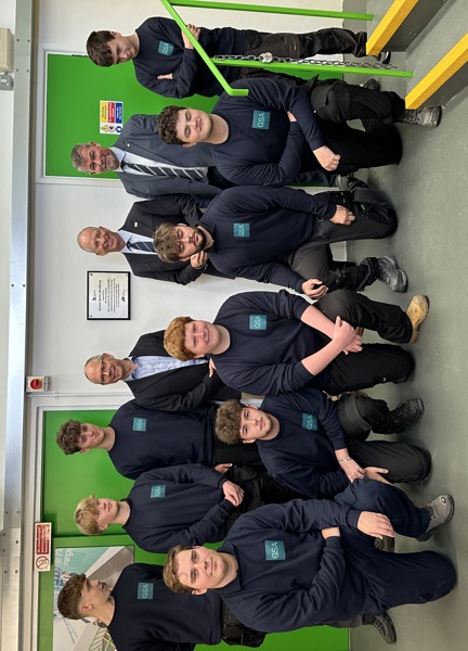 A group of Green Skills Academy apprentices wearing their Green Skills uniform together indoors in front of a plaque on the wall. Standing with them, wearing suits, are Chris Amber - JE CEO, Lieutenant Governor Vice Admiral Sir Jerry Kyd KBE, and Lance Chatfield - Principal of Highlands College. The room has green doors and a white wall.