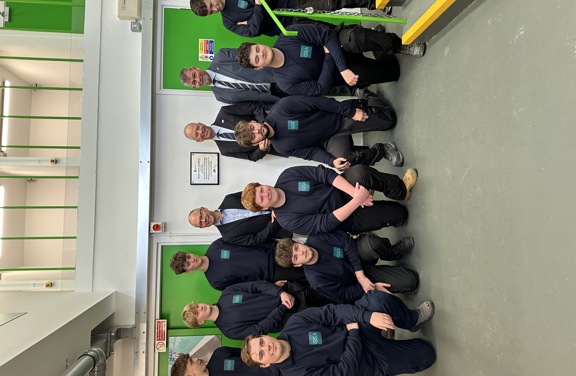 A group of Green Skills Academy apprentices wearing their Green Skills uniform together indoors in front of a plaque on the wall. Standing with them, wearing suits, are Chris Amber - JE CEO, Lieutenant Governor Vice Admiral Sir Jerry Kyd KBE, and Lance Chatfield - Principal of Highlands College. The room has green doors and a white wall.