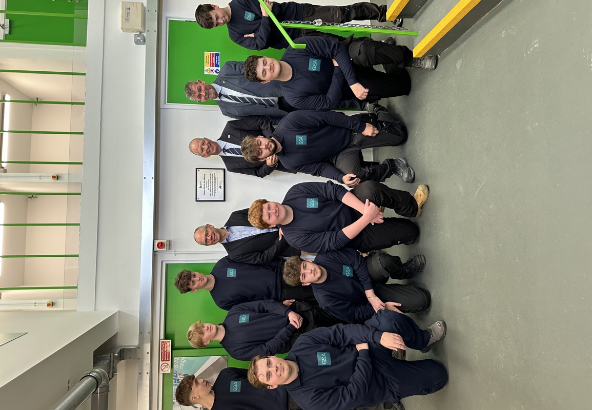 A group of Green Skills Academy apprentices wearing their Green Skills uniform together indoors in front of a plaque on the wall. Standing with them, wearing suits, are Chris Amber - JE CEO, Lieutenant Governor Vice Admiral Sir Jerry Kyd KBE, and Lance Chatfield - Principal of Highlands College. The room has green doors and a white wall.
