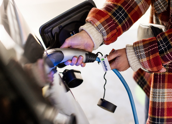 A close up photo of a person in a red checked jacket plugging in a charging cble to their electric vehicle