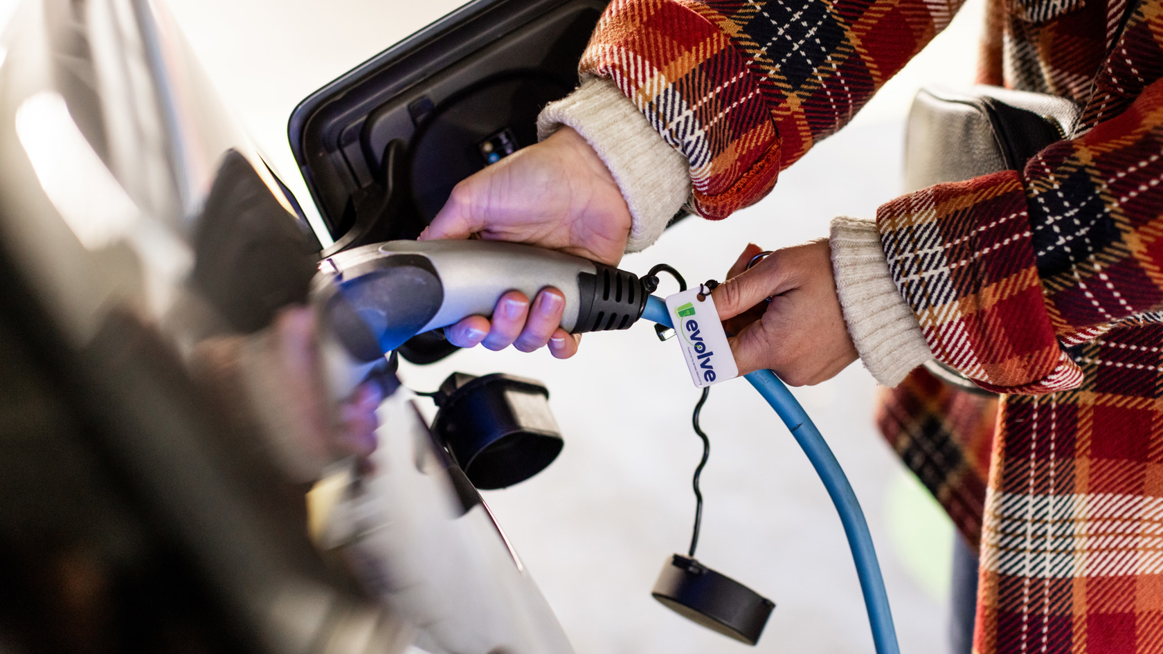 A close up photo of a person in a red checked jacket plugging in a charging cble to their electric vehicle