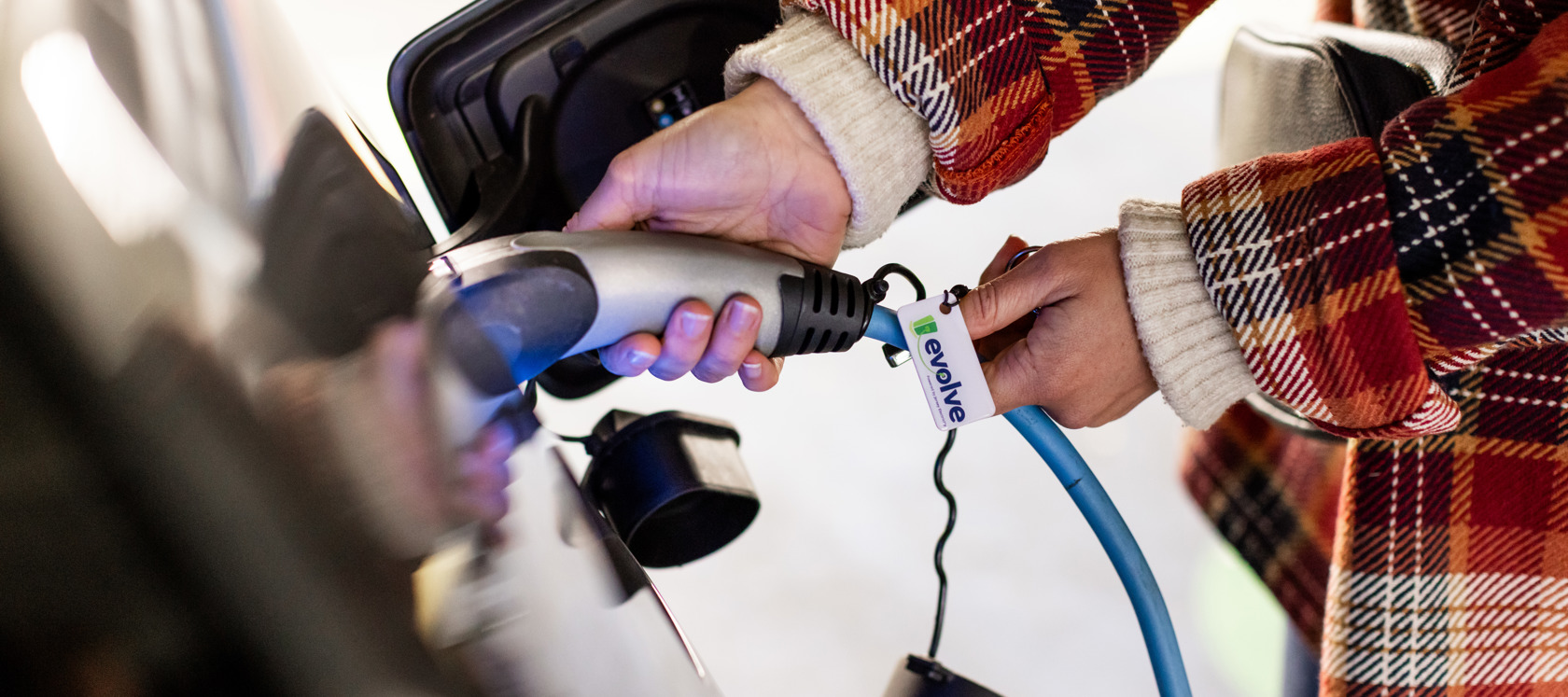 A close up photo of a person in a red checked jacket plugging in a charging cble to their electric vehicle