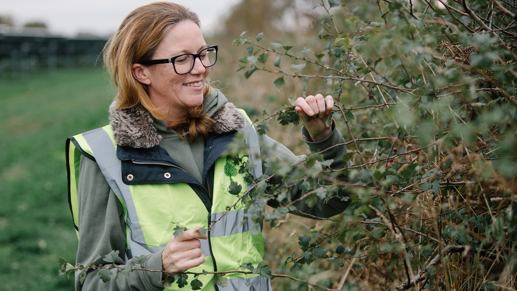 Alex Morel standing in a field wearing a high vis vest and smiling which she inspects the hedgerows