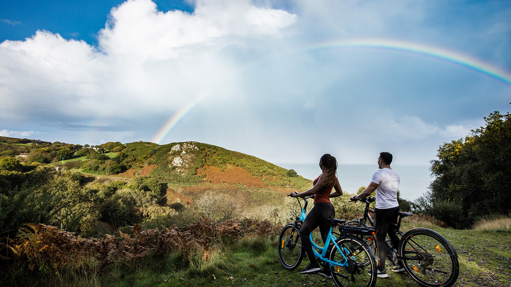 A man and a women look at a rainbow while riding electric bicycles.