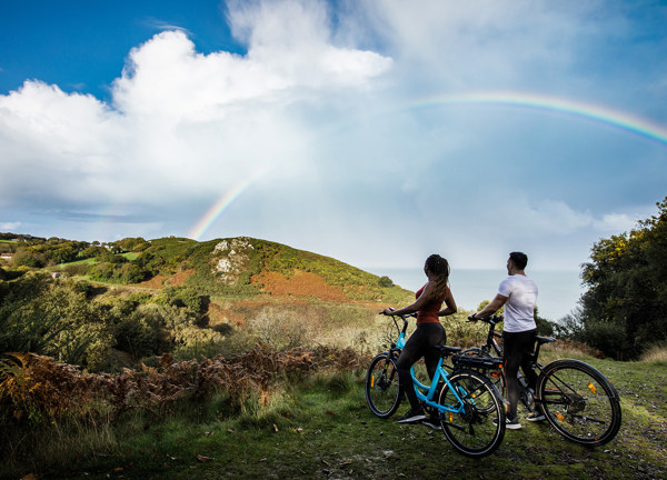 A man and a women look at a rainbow while riding electric bicycles.